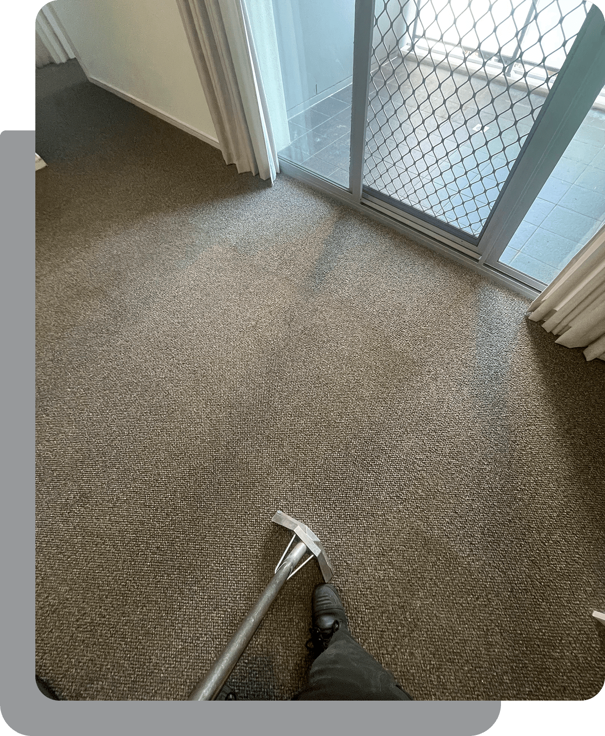 A person operates a carpet cleaning tool on a brown carpet near a glass door with a screen.